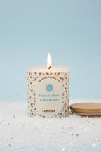 Candela artigianale Luminah Cime Innevate accesa, in vetro satinato bianco, su sfondo azzurro con neve decorativa e coperchio in bamboo.