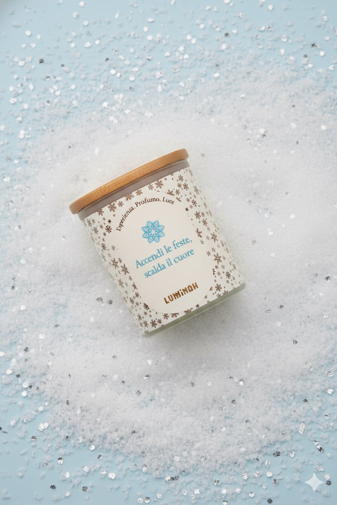 Candela artigianale Luminah Cime Innevate in vetro satinato con coperchio in bamboo, poggiata su neve decorativa e sfondo azzurro.
